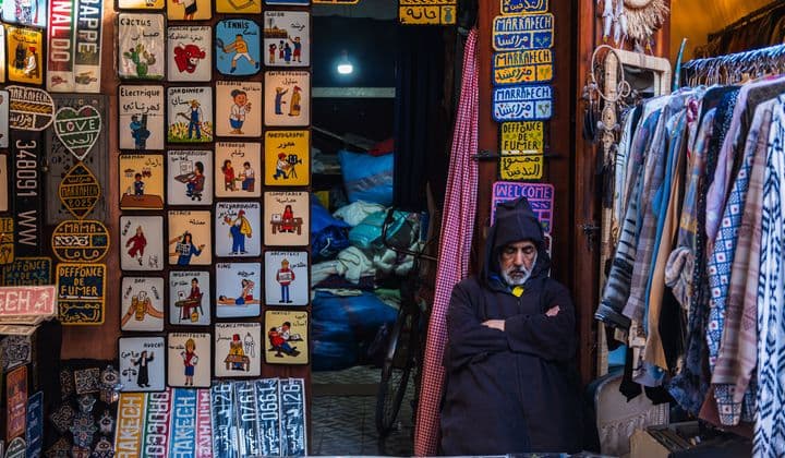 A vendor in Marrakech souk, surrounded by vibrant signs and garments, embodies local culture.